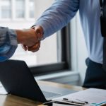 Lease Contract - Crop unrecognizable coworkers in formal wear standing at table with laptop and documents while greeting each other before meeting